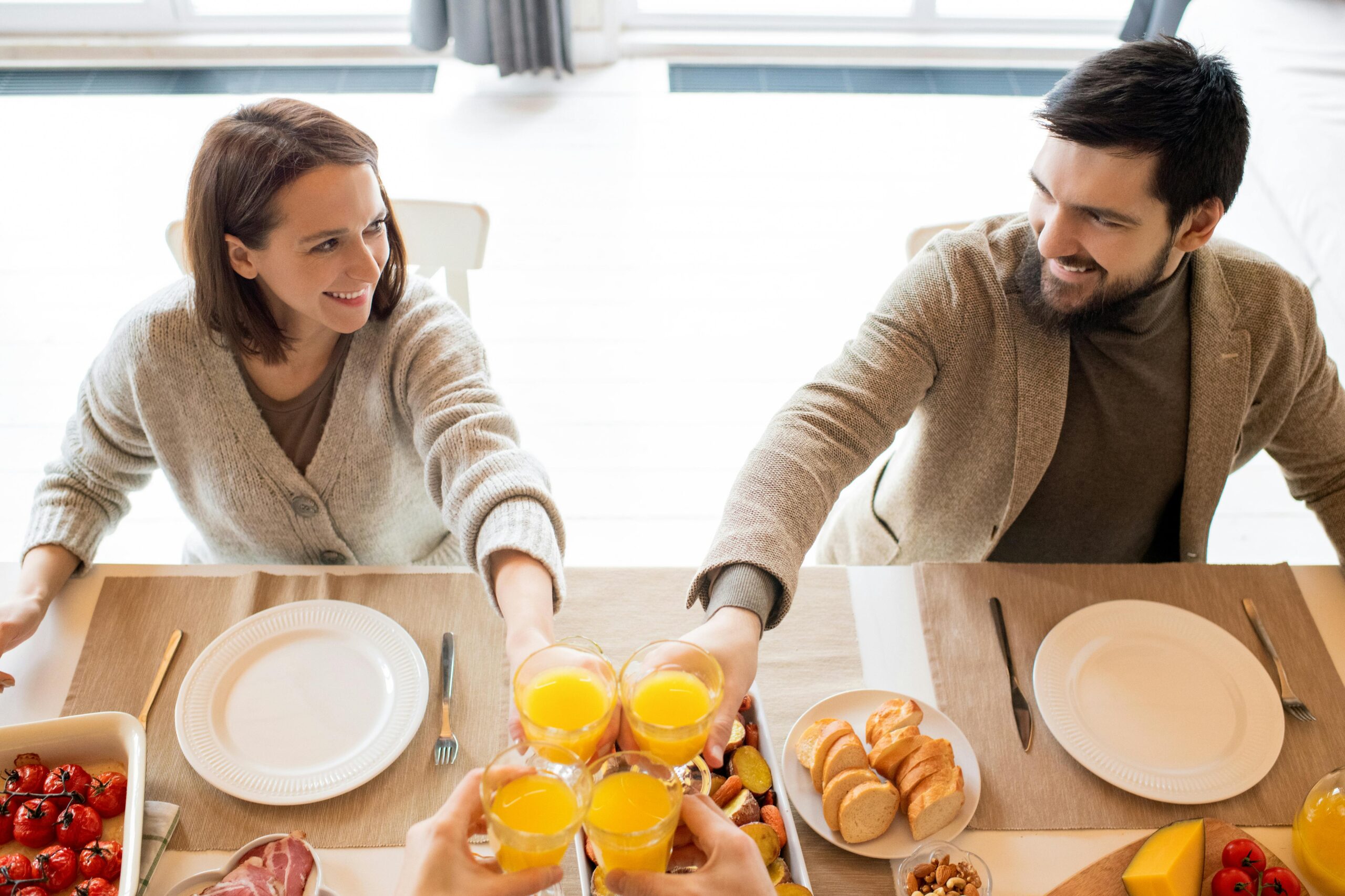 A joyful couple shares a toast with orange juice at a sunny breakfast table indoors.